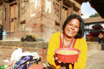 Smiling woman in traditional clothing holding a bowl of colourful grains in a sunlit courtyard – reflecting joy, generosity, and the everyday strength rooted in cultural pride.