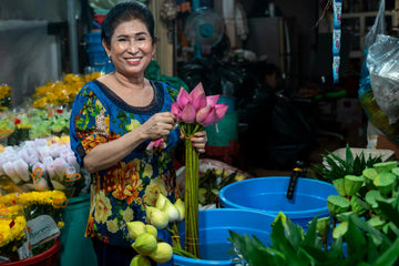 Smiling woman arranging lotus flowers at a vibrant flower stall, surrounded by colourful blooms and blue buckets – celebrating care, craft, and the joyful rhythm of daily work.