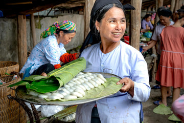 Smiling woman holding a tray of traditional rice cakes wrapped in green leaves at a local market – showcasing cultural pride, handcrafted care, and the strength of community tradition.