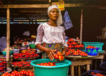 Woman sorting tomatoes at a vibrant outdoor market stall, surrounded by colourful baskets and produce – representing strength, independence, and the dignity of daily work.