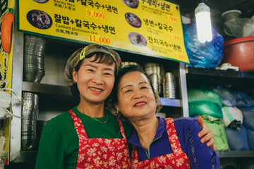 Two smiling women wearing floral aprons standing in their market food stall beneath a bright Korean menu – capturing partnership, pride, and everyday strength in local culinary culture.