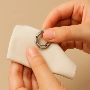 A hand gently wiping an EQUØRA stainless steel ring with a soft beige microfibre cloth on a matching neutral background.