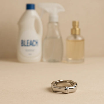 An EQUØRA stainless steel ring in sharp focus on a beige surface, with blurred bottles of bleach, a household cleaner spray and a perfume atomiser in the background.