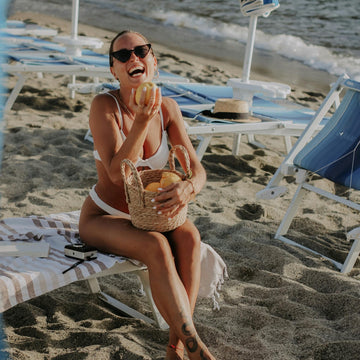 Woman in a white bikini laughing with a basket of fruit by the shore – radiating body confidence, lightness, and self-nourishment.