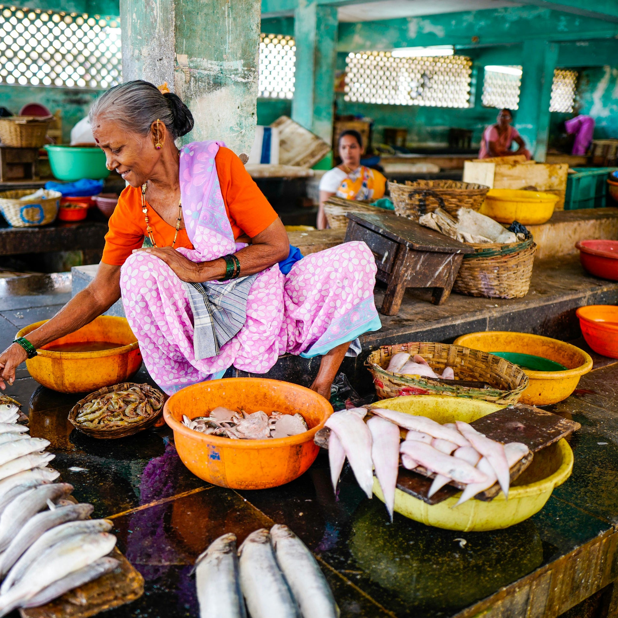 Elderly woman selling fresh fish at a colourful local market, seated among baskets and bowls – honouring labour, dignity, and the everyday strength of women in community trade.