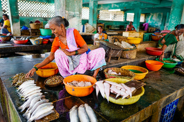 Elderly woman selling fresh fish at a colourful local market, seated among baskets and bowls – honouring labour, dignity, and the everyday strength of women in community trade.
