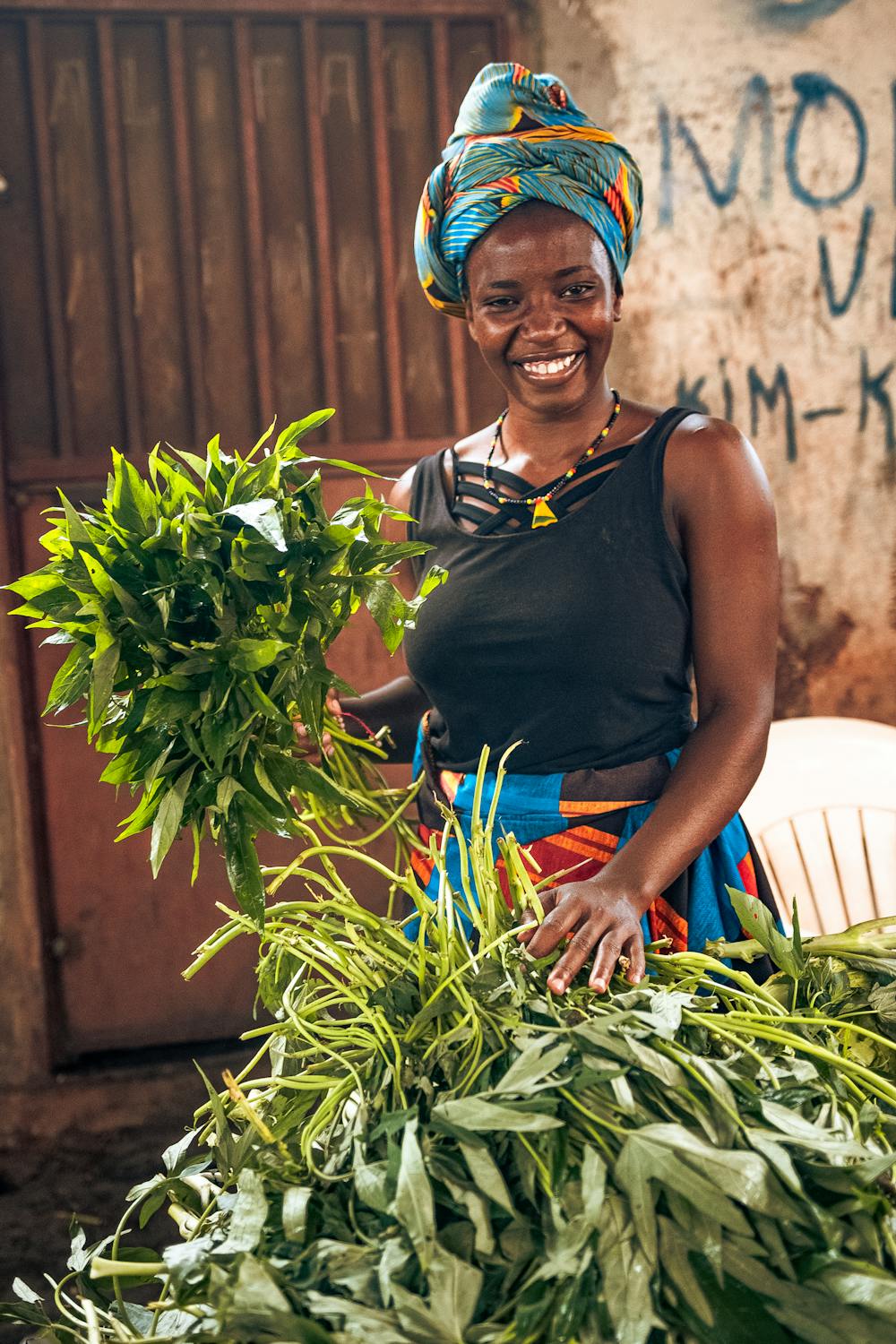 Smiling woman in colourful headwrap and beaded jewellery holding freshly harvested greens at a local market – celebrating strength, joy, and everyday resilience in motion.