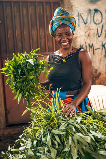 Smiling woman in colourful headwrap and beaded jewellery holding freshly harvested greens at a local market – celebrating strength, joy, and everyday resilience in motion.