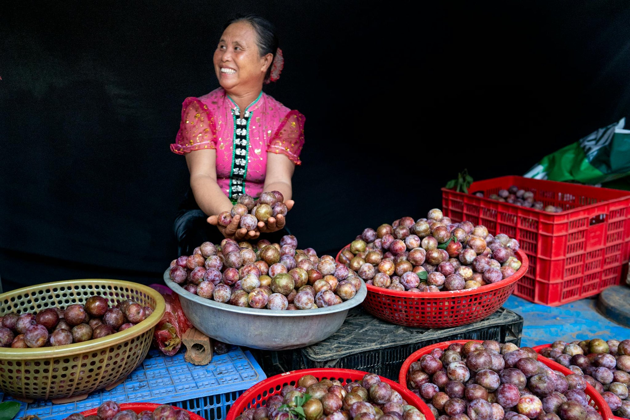 Smiling woman in vibrant traditional clothing holding fresh plums at a market stall surrounded by full baskets – celebrating abundance, joyful labour, and cultural heritage through community trade.