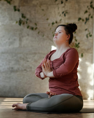 Young woman practising seated yoga meditation with hands in prayer position, wearing a maroon top and grey leggings in a calm, sunlit indoor space with a rustic wall and leafy vines in the background.