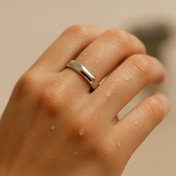 Close-up of a fair-skinned hand wearing an EQUØRA 316L stainless steel ring, with water droplets on the skin, set against a neutral beige background.