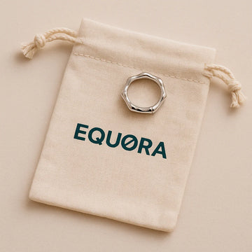 A natural cotton drawstring pouch printed with a dark green EQUØRA logo, the top gently cinched and a stainless steel ring peeking out, on a soft beige surface.