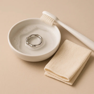 A white ceramic bowl of warm, soapy water holding an EQUØRA stainless steel ring, a soft-bristled toothbrush resting on the rim and a folded beige microfibre cloth beside it, all on a neutral beige background.