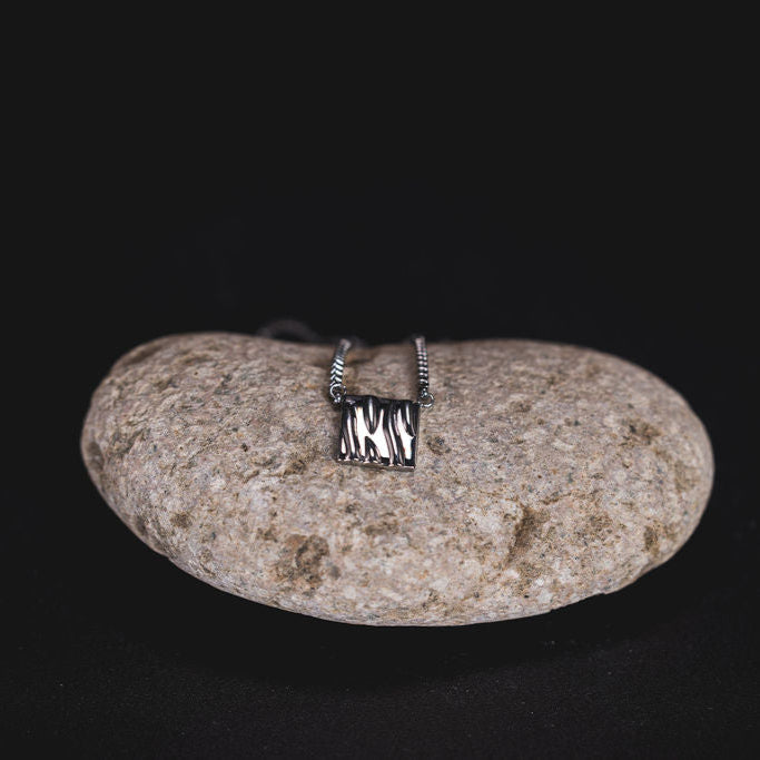 EQUØRA Coast Necklace with textured square pendant, elegantly displayed on a natural stone against a black background — a modern statement piece inspired by organic forms.