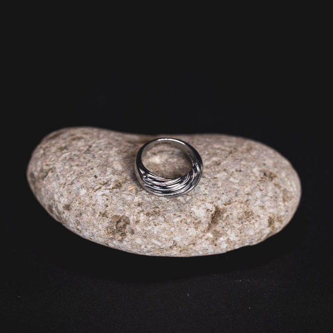 EQUØRA Current Ring — sculptural stainless steel ring with fluid, wave-like detailing, styled on a natural stone surface for a refined and organic look. Ideal for statement styling.