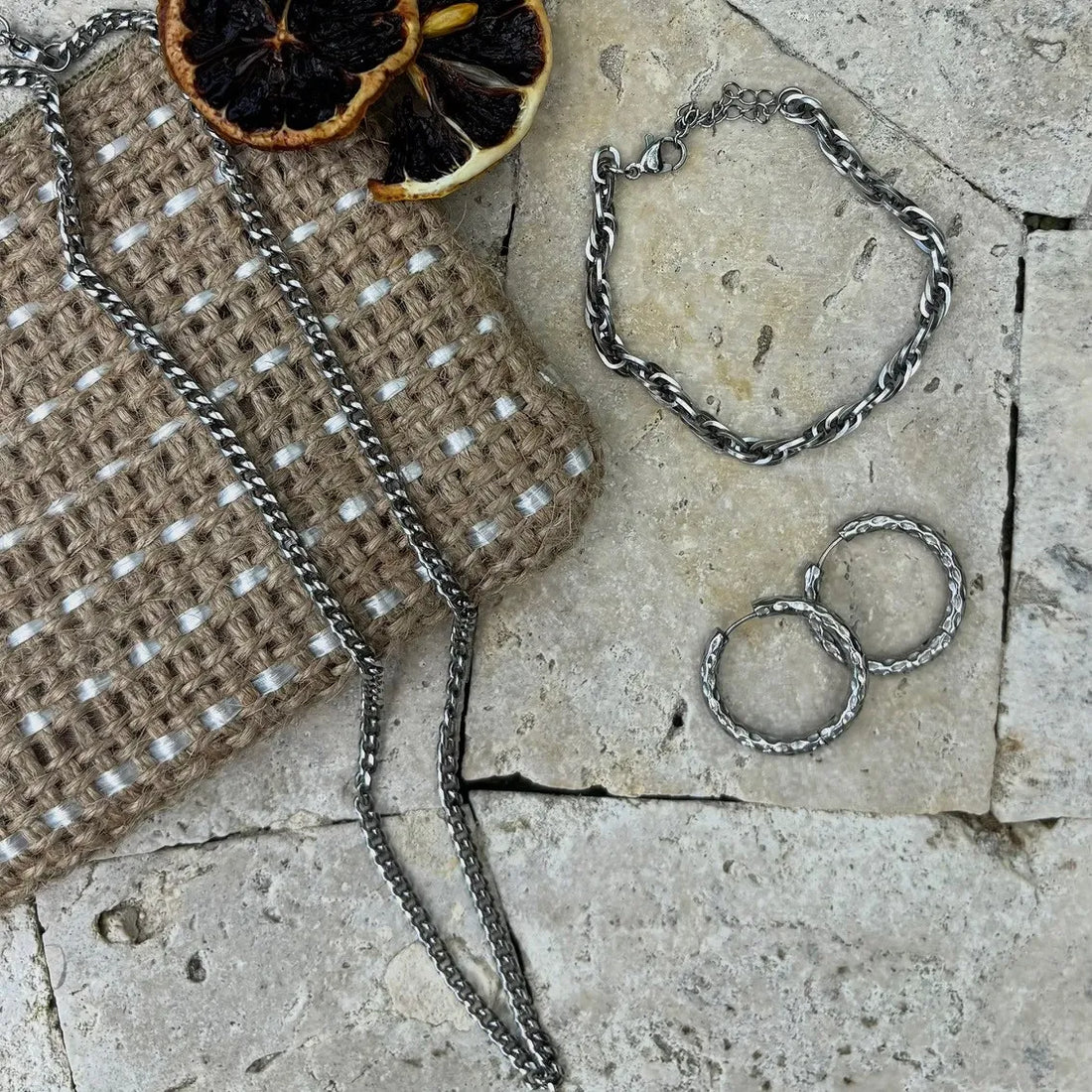 Flat lay of silver twisted hoop earrings, a matching chain bracelet, and a woven pouch on a stone surface, with dried citrus slices for styling.