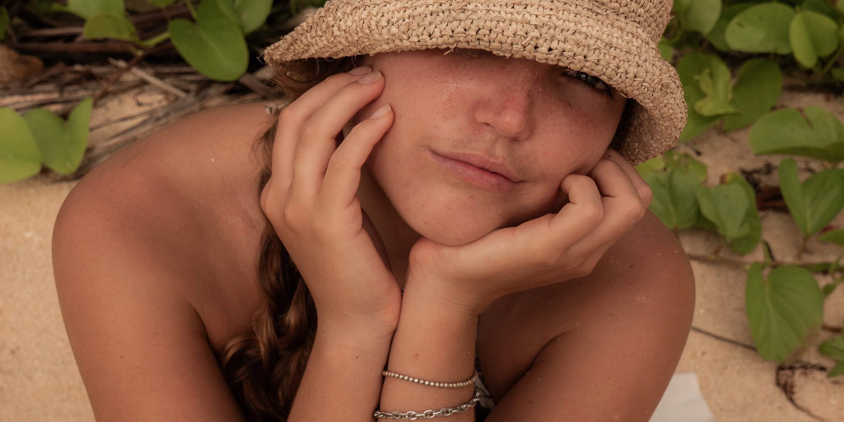 Woman in a woven sun hat sitting on the sand with hands on her face, wearing silver bracelets and surrounded by greenery – reflecting calm, introspection, and the essence of slow, intentional living.