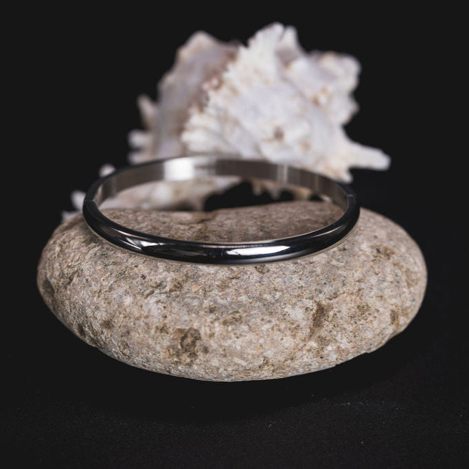 EQUØRA Frame Bracelet — sleek, polished silver bangle with a modern minimalist design, elegantly placed on a smooth natural stone with a coral accent in the background.