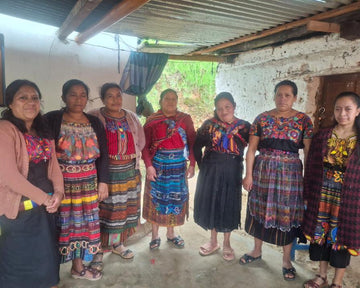 Seven women from the Guadalupe Rancho Group in Totonicapan, Guatemala, stand together in traditional woven garments, smiling in a modest shelter. Supported by EQUØRA and Kiva.org through Friendship Bridge, they receive microloans and business training to grow their enterprises and improve family wellbeing.