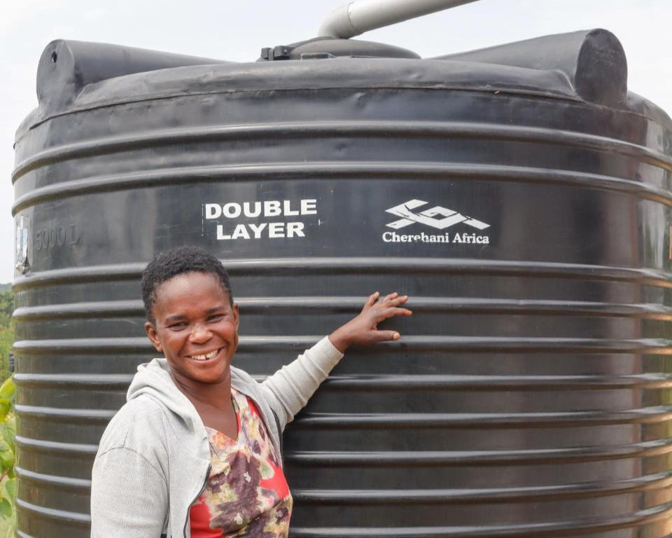 Smiling woman in rural Kenya stands beside a large black water tank labelled "Double Layer" and "Cherehani Africa", supported by EQUØRA and Kiva.org to help provide clean water and empower women entrepreneurs through microfinance solutions.