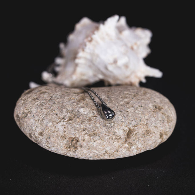 EQUØRA Mist Necklace featuring a smooth teardrop pendant on a fine chain, styled atop a natural stone with a shell backdrop — minimalist jewellery inspired by nature's quiet elegance.