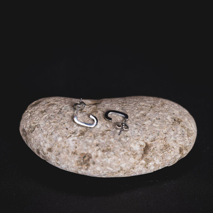 EQUØRA Nivra Earrings — asymmetrical stainless steel hoops with modern geometric design, displayed on a smooth stone. Unique minimalist earrings for everyday style.