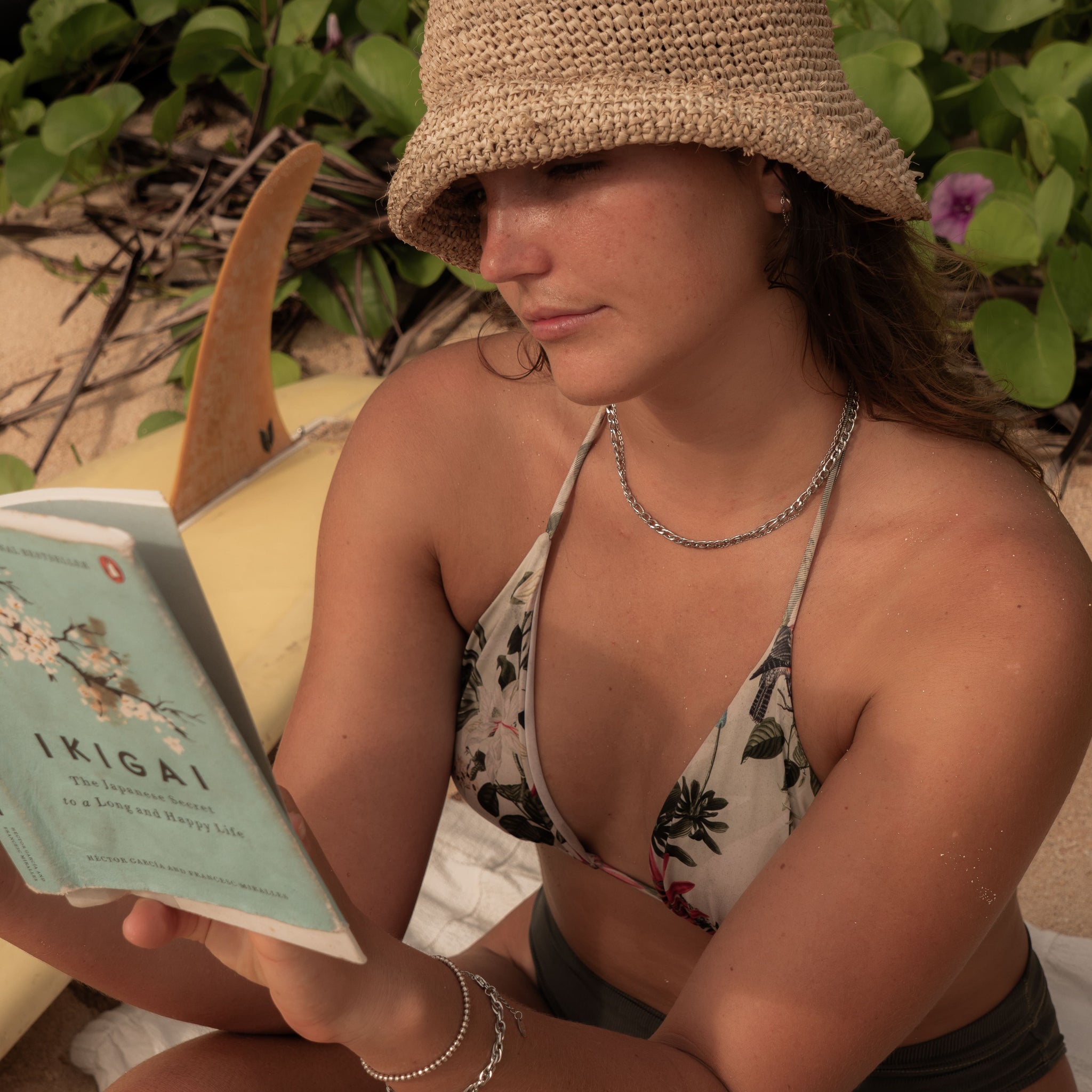 Woman in silver jewellery and floral swimwear reading 'Ikigai' on the beach, surfboard beside her – capturing a moment of calm intention, inner strength, and fluid living.