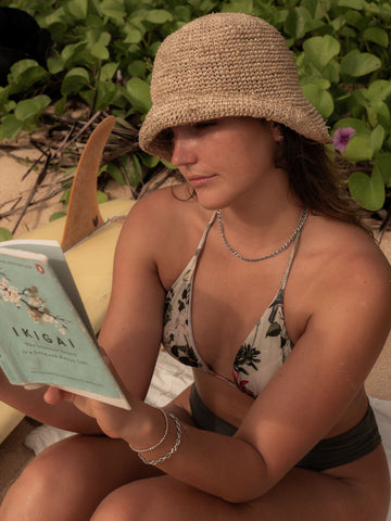 Woman in silver jewellery and floral swimwear reading 'Ikigai' on the beach, surfboard beside her – capturing a moment of calm intention, inner strength, and fluid living.