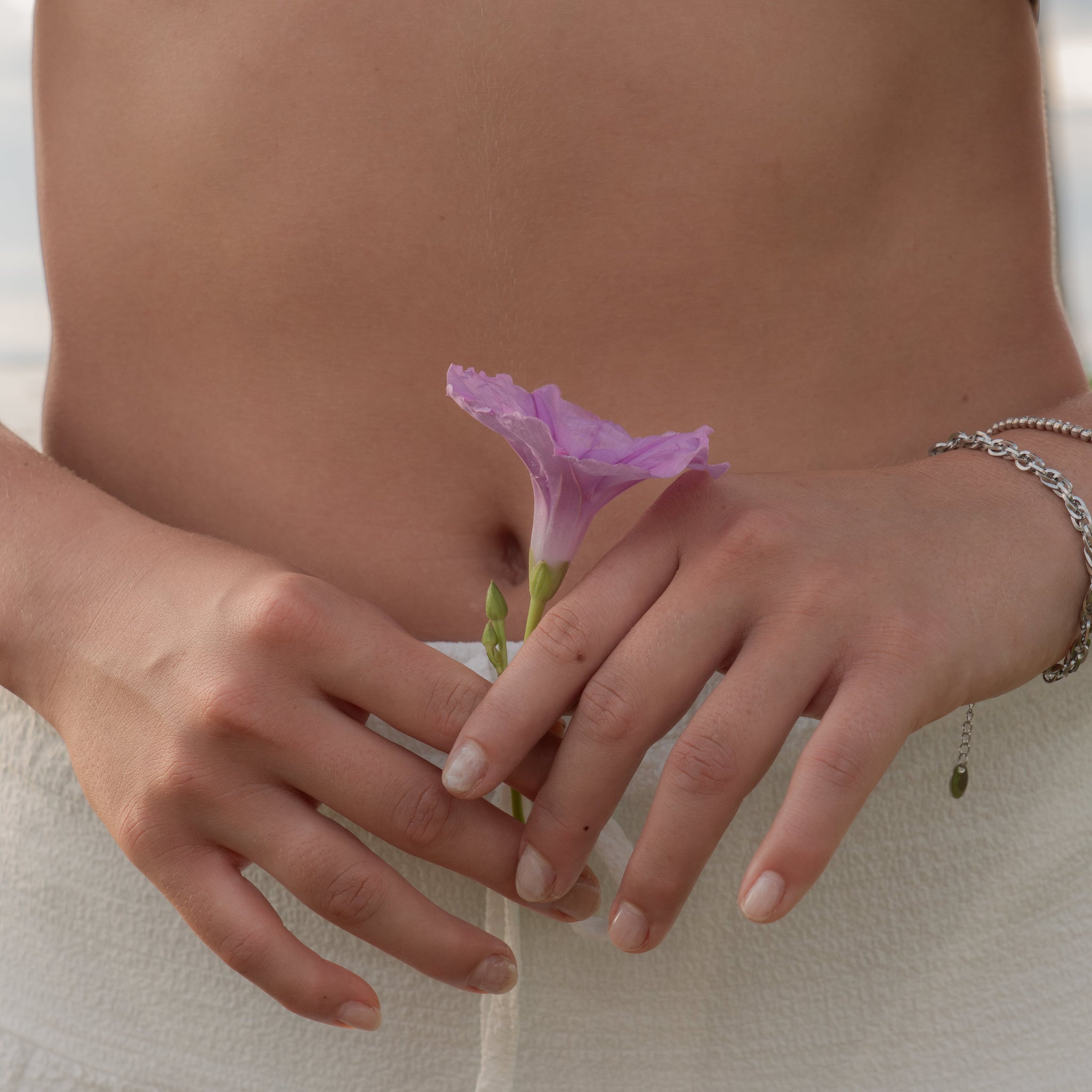 Close-up of a woman’s hands gently holding a purple flower against her body, wearing silver jewellery – capturing EQUØRA’s story of softness, strength, and quiet intention in motion.