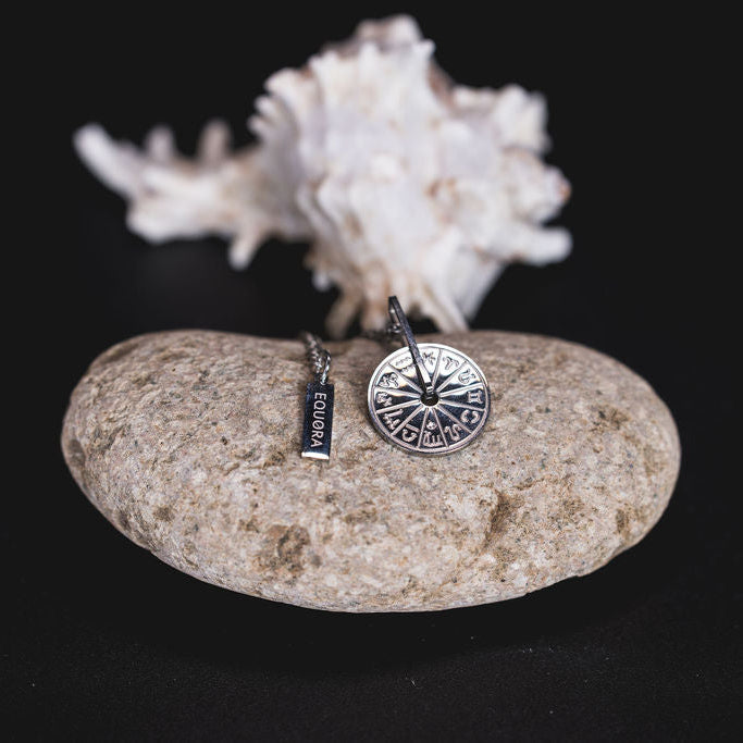 EQUØRA Outshine Fate Necklace with a detailed compass pendant and engraved cardinal points, styled on a smooth stone and seashell — symbolic stainless steel jewellery for direction and inner power.
