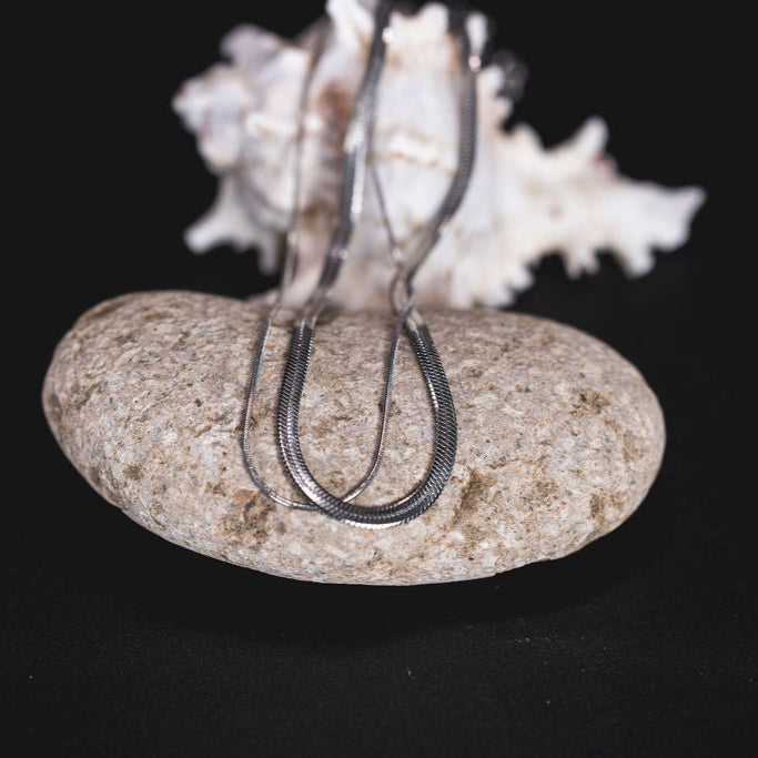 EQUØRA Serpentis Necklace with layered snake chain design, elegantly draped over a smooth beach stone and seashell — minimalist stainless steel jewellery with coastal influence.