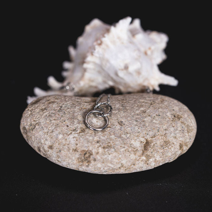 EQUØRA Unity Necklace with two interlocking rings symbolising connection and strength, displayed on a natural stone with a white seashell backdrop — timeless minimalist jewellery design.