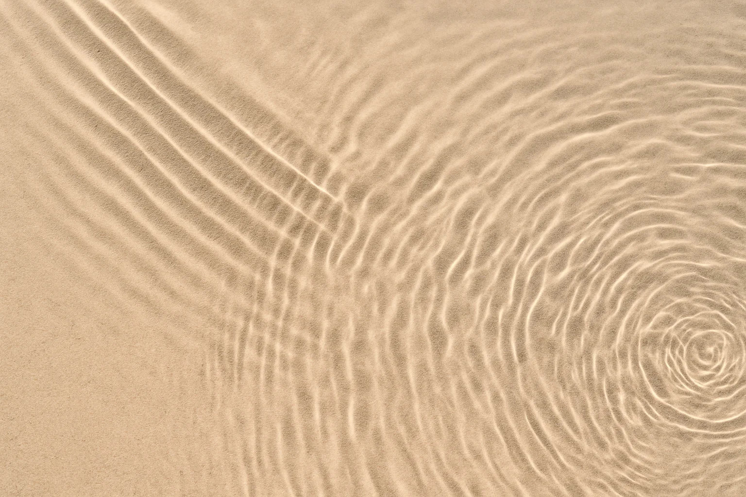 Overhead view of soft water ripples forming concentric patterns on golden beach sand – symbolising calm, clarity, and the quiet strength behind every EQUØRA piece.